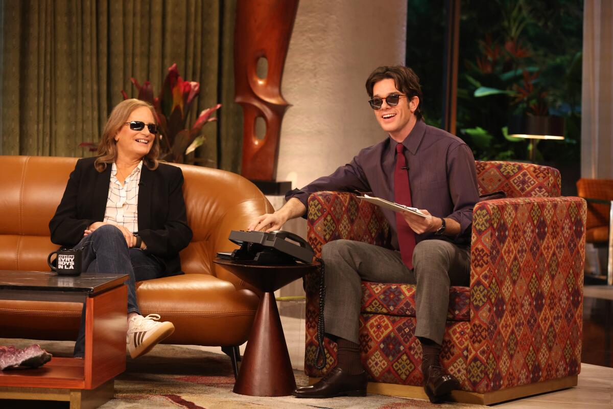 Two people sitting on a TV show set, both wearing dark sunglasses