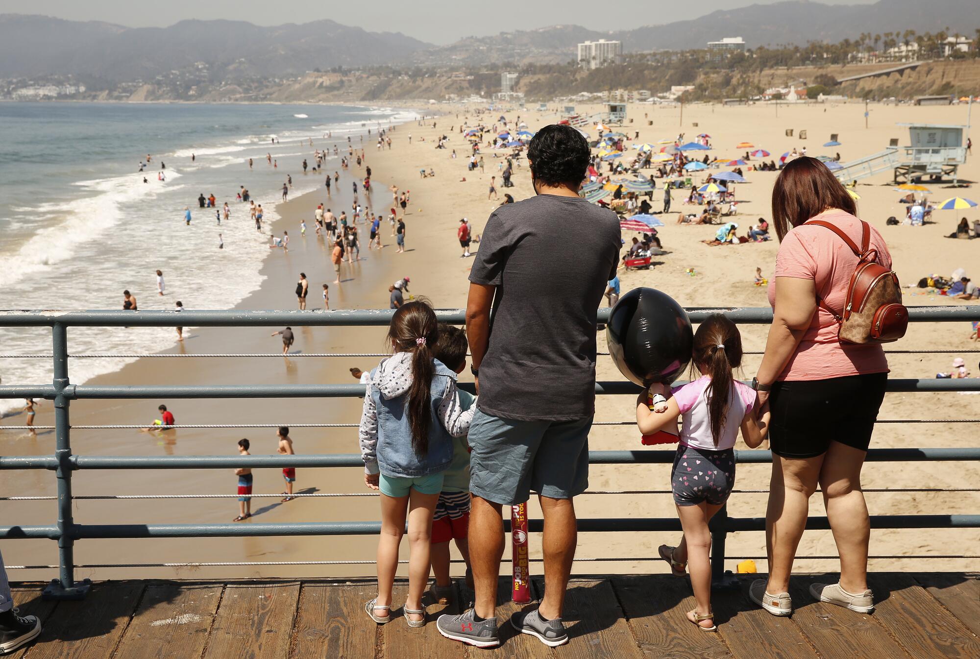 A man, woman and three small children look from a pier over a busy beach.