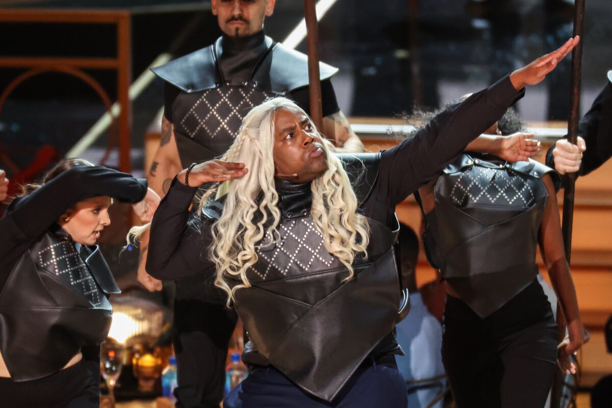 A man in a blond wig and black body suit dances with others onstage 