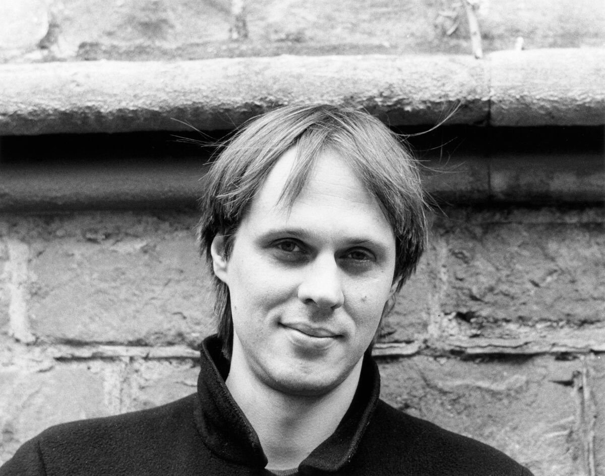 Close-up black-and-white photo of a young Tom Verlaine standing in front of a concrete wall.
