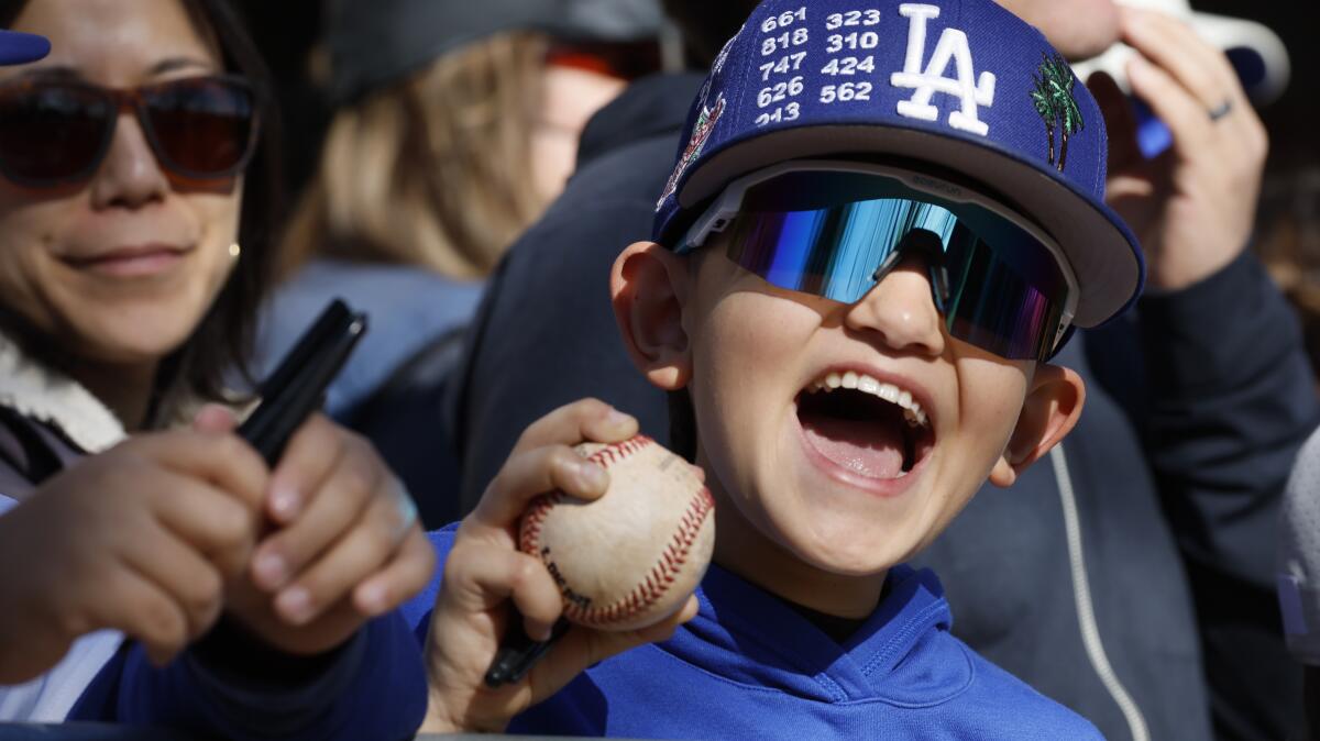 A little boy wearing a Dodgers cap and sunglasses smiles while holding a baseball