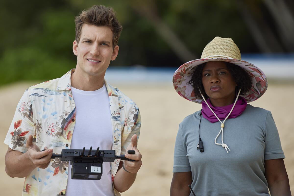 A man holding a drone remote control stands on a beach next to a woman wearing a straw hat.