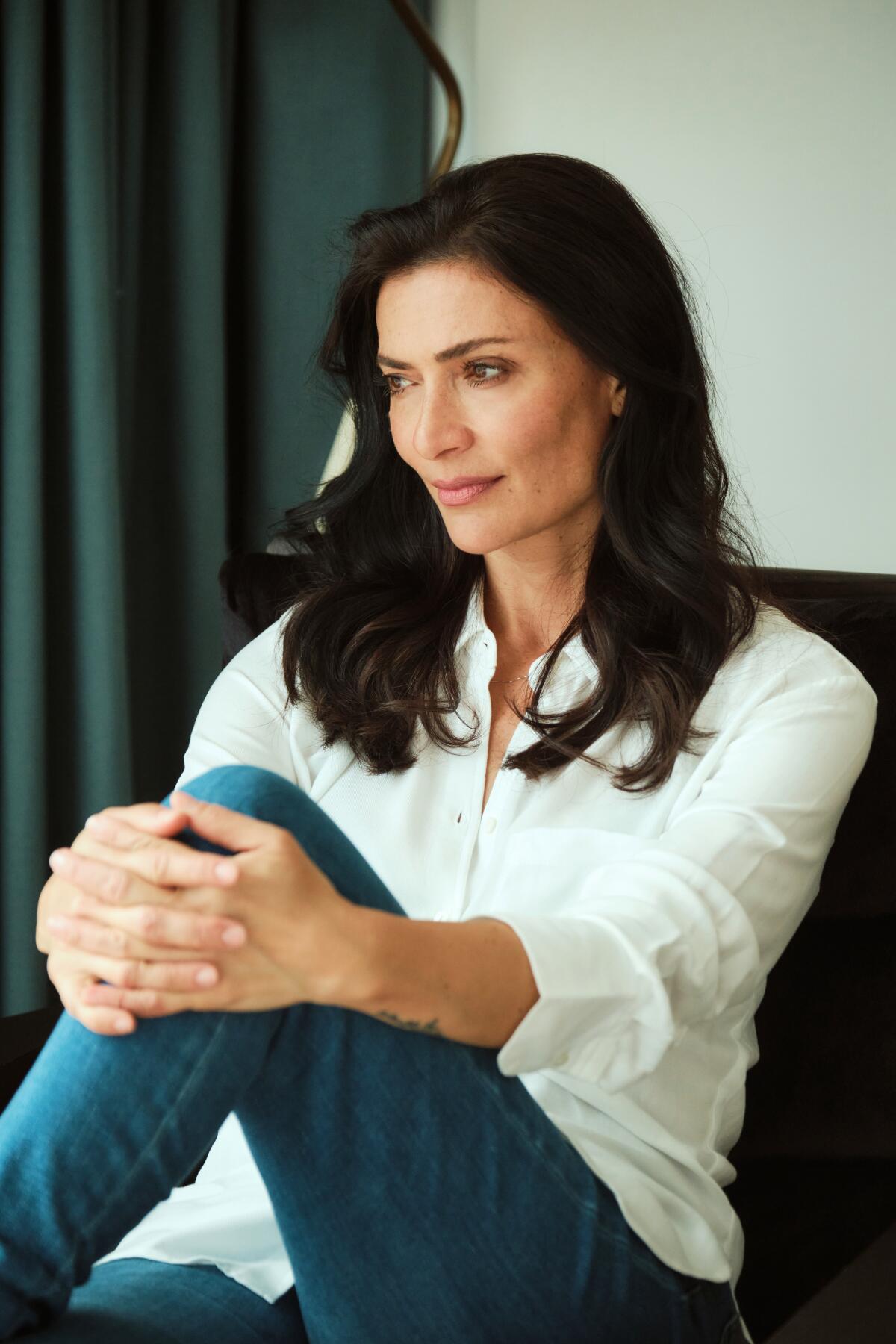 Ana Maria Orozco in a white shirt and blue jeans sitting with her hands wrapped around her knee.
