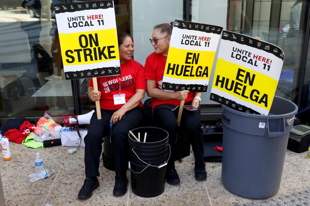 Two women sit outside of a building, both holding pickets signs while looking at each other.