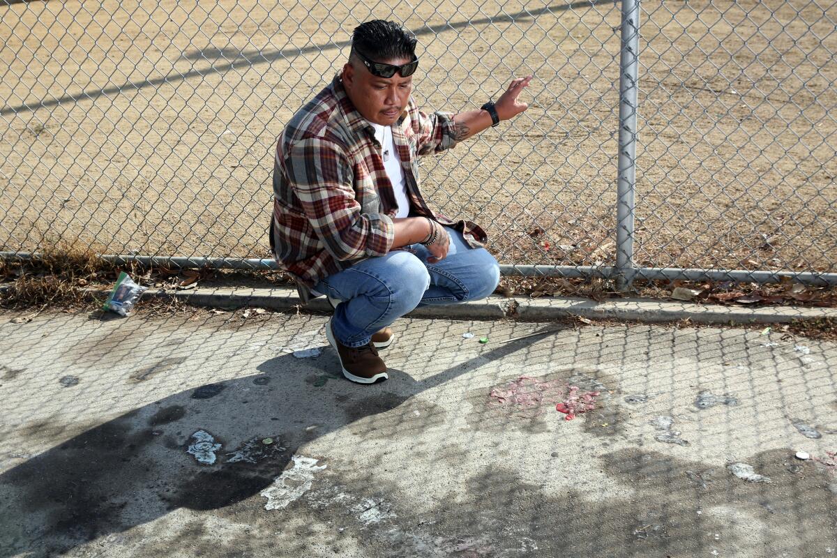 A man kneels on a patch of sidewalk with his hand on a chain-link fence