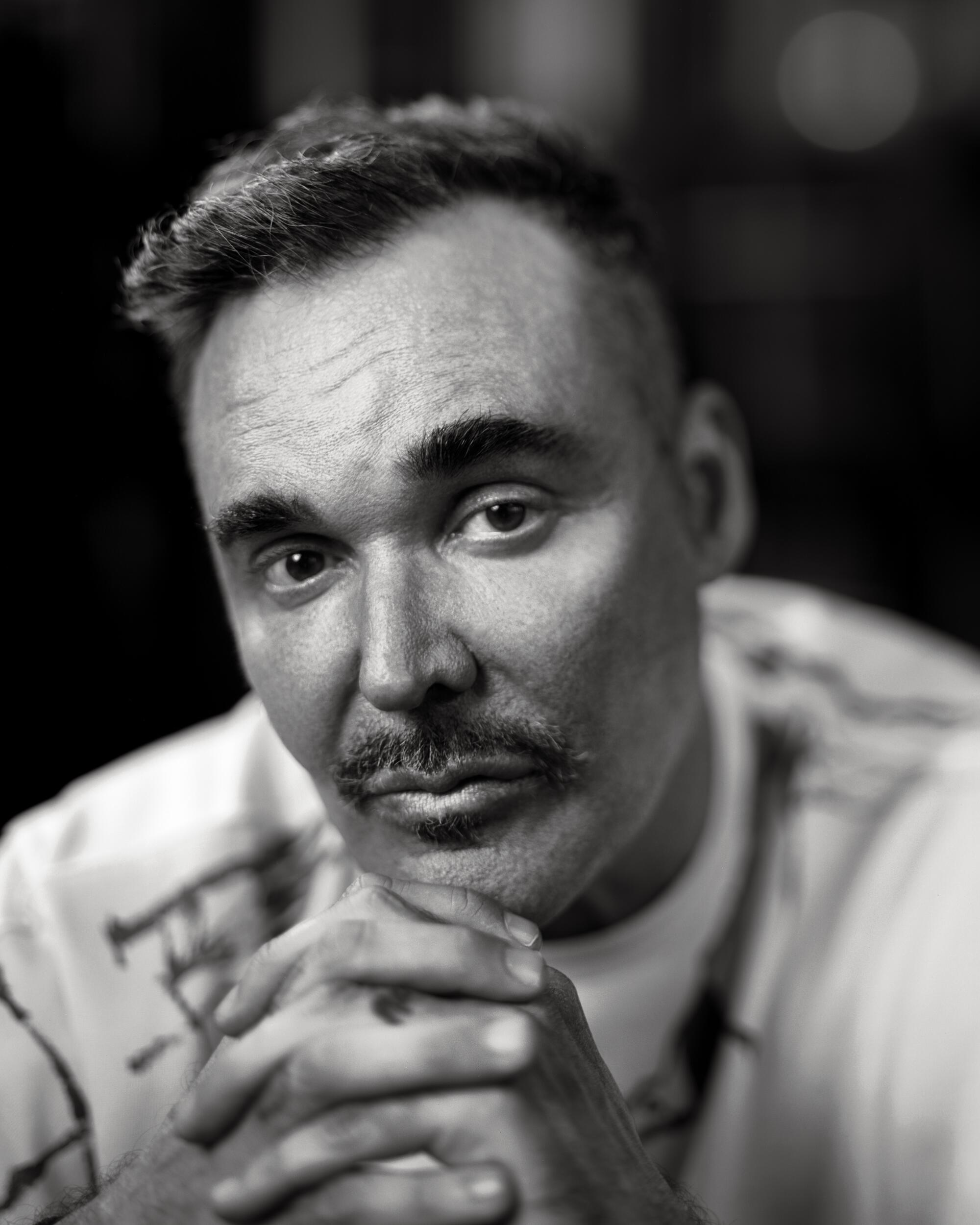 A black-and-white close-up of David LaChapelle with his hands crossed under his chin.