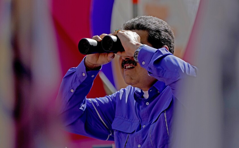 A man with dark hair and mustache, wearing a purple shirt, looks through binoculars 