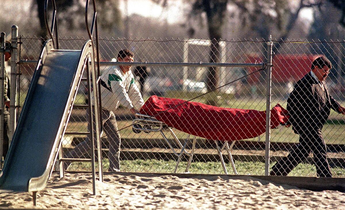 Two men wheel a body in a red bag on a gurney past a playground