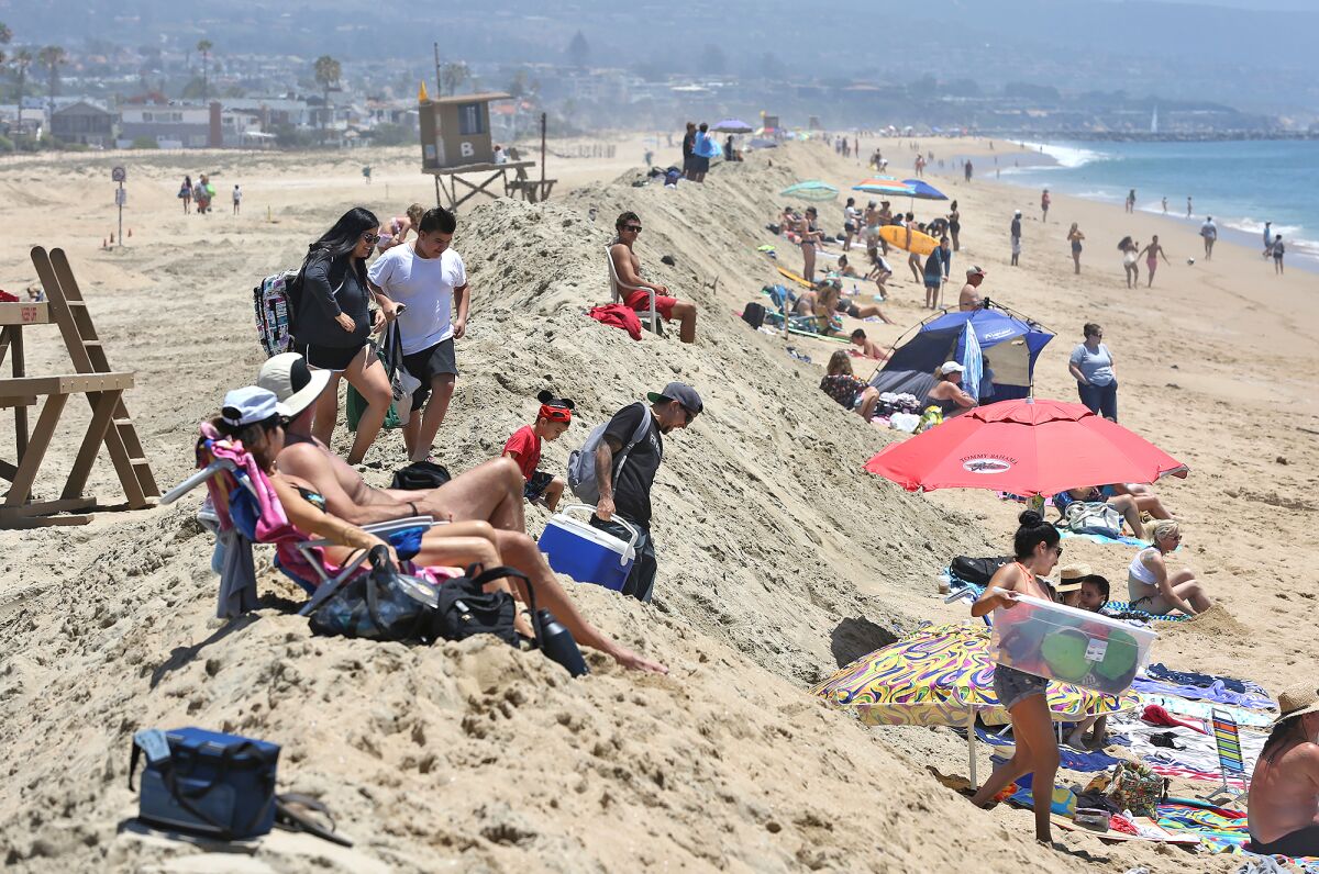 Beachgoers climb and sit on top of a sand berm that was rebuilt to hold off big waves and high tides at Balboa Pier.