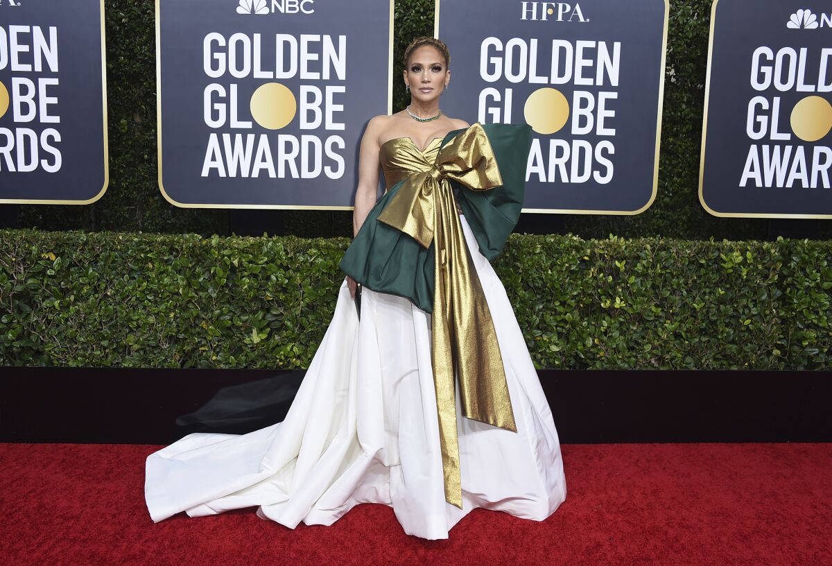 Jennifer Lopez stands on a red carpet in a formal green and white gown with voluminous skirt and oversized gold bow.