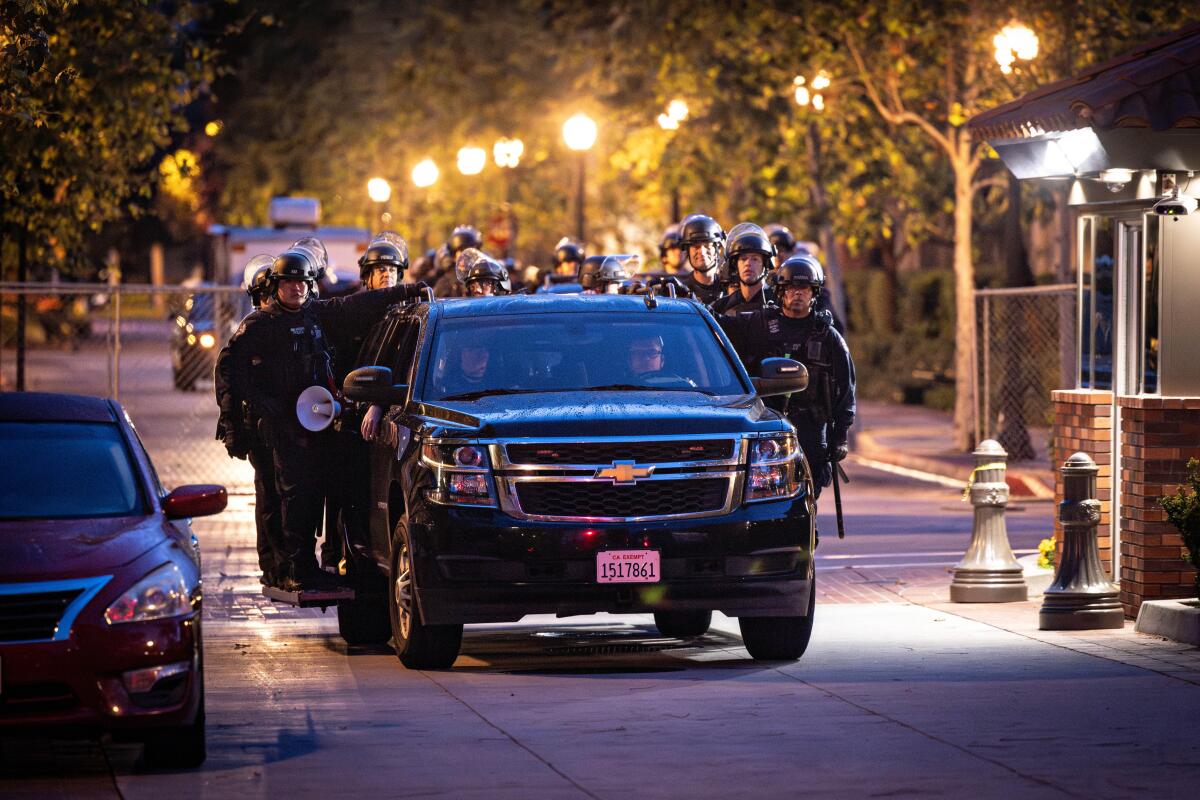 LAPD officers in riot gear exit USC behind a vehicle at night.