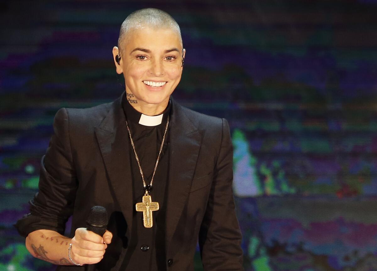Sinead O'Connor smiling while on stage, holding a mic in one hand, wearing a black suit, priest's collar and golden cross