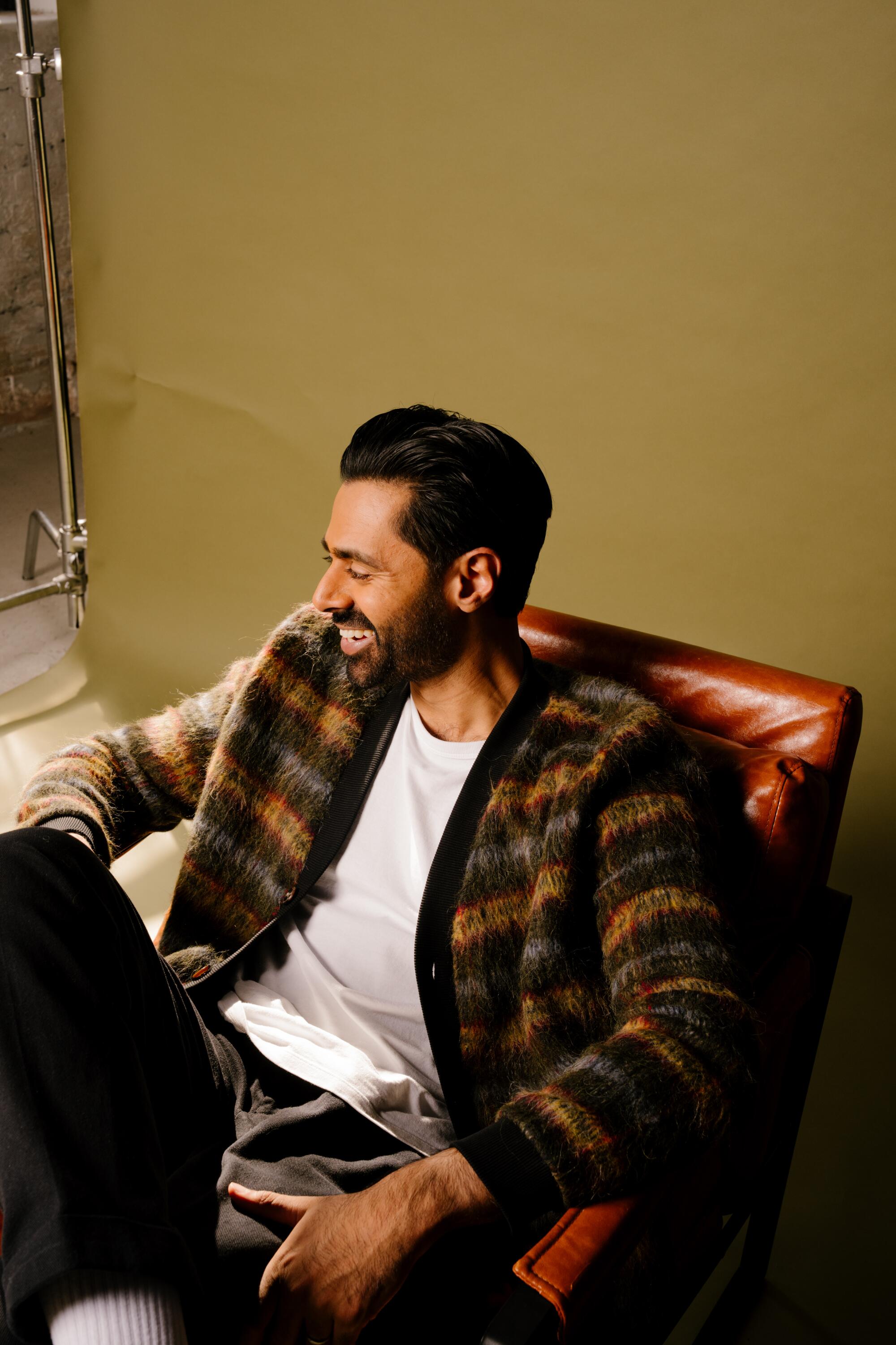 A man in a patterned sweater, white T-shirt and dark pants sits in a red chair laughing, with his face turned away.
