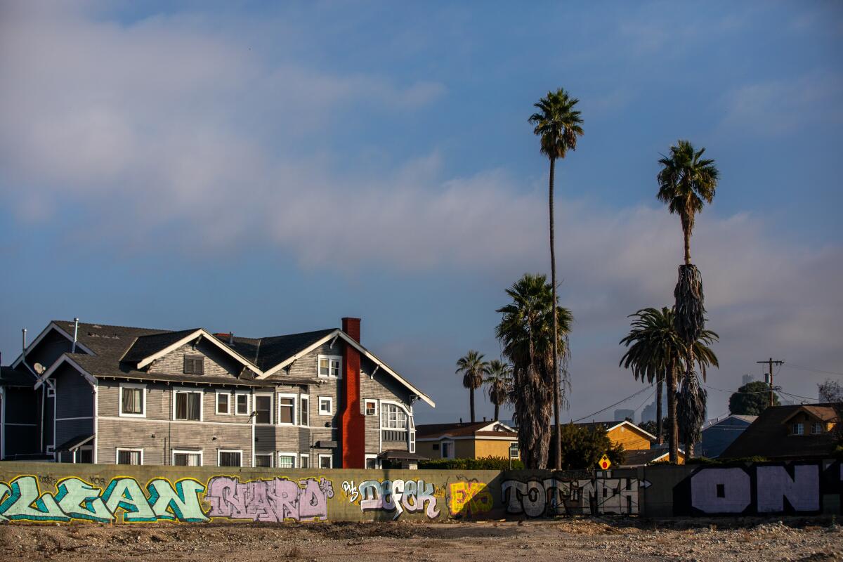 An apartment complex and palm trees stand behind a dirt lot enclosed by a wall covered in graffiti. 