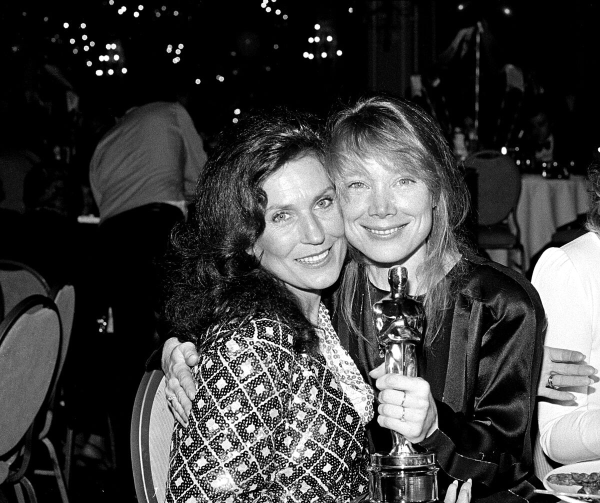 Two women pose for a photo. The woman on the right is holding an Oscar.