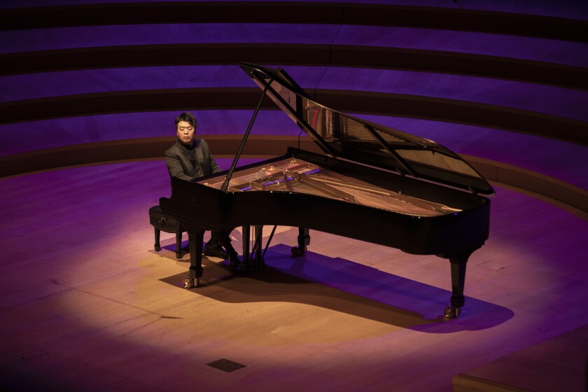 A man in a dark gray suit seated at a grand piano under a spotlight.
