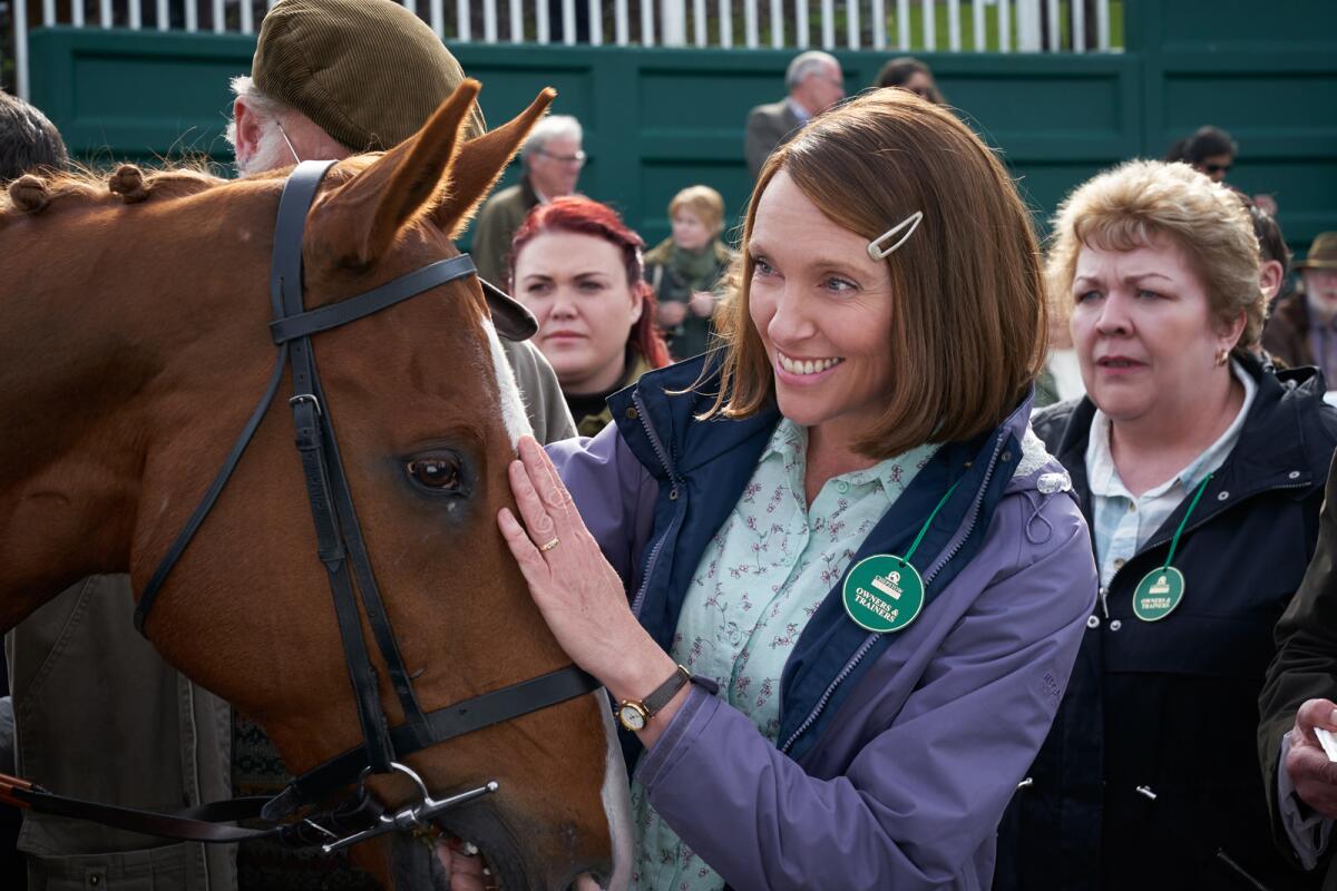 Toni Collette, amid a crowd, smiles as she touches a horse on the head. 
