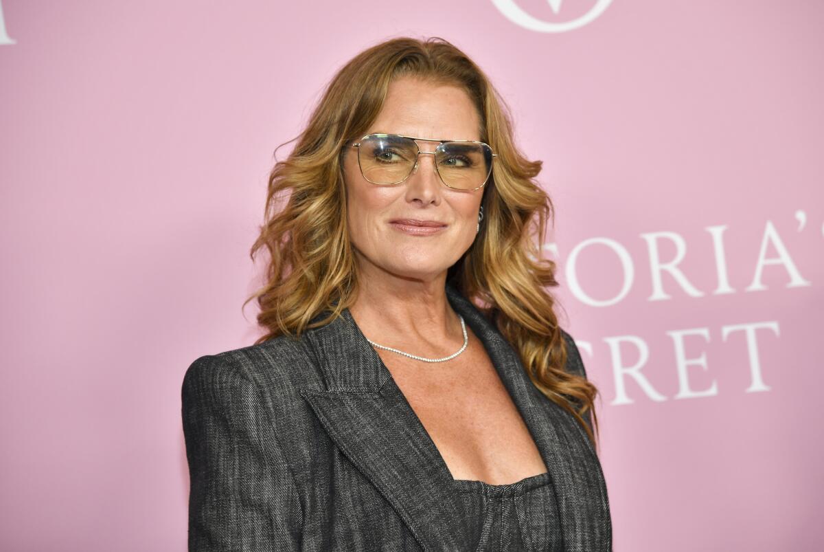 Brooke Shields wears a gray pantsuit and glasses as she poses for photos in front of a pink background