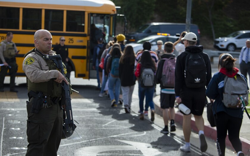 A sheriff's deputy, holding a gun pointed down, looks on as students board buses en route to a family reunification area after Thursday's deadly shooting at Saugus High School in Santa Clarita.