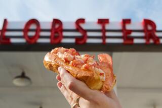 A hand holds a massive lobster roll in front of the vintage gas station's red "LOBSTER" signage.
