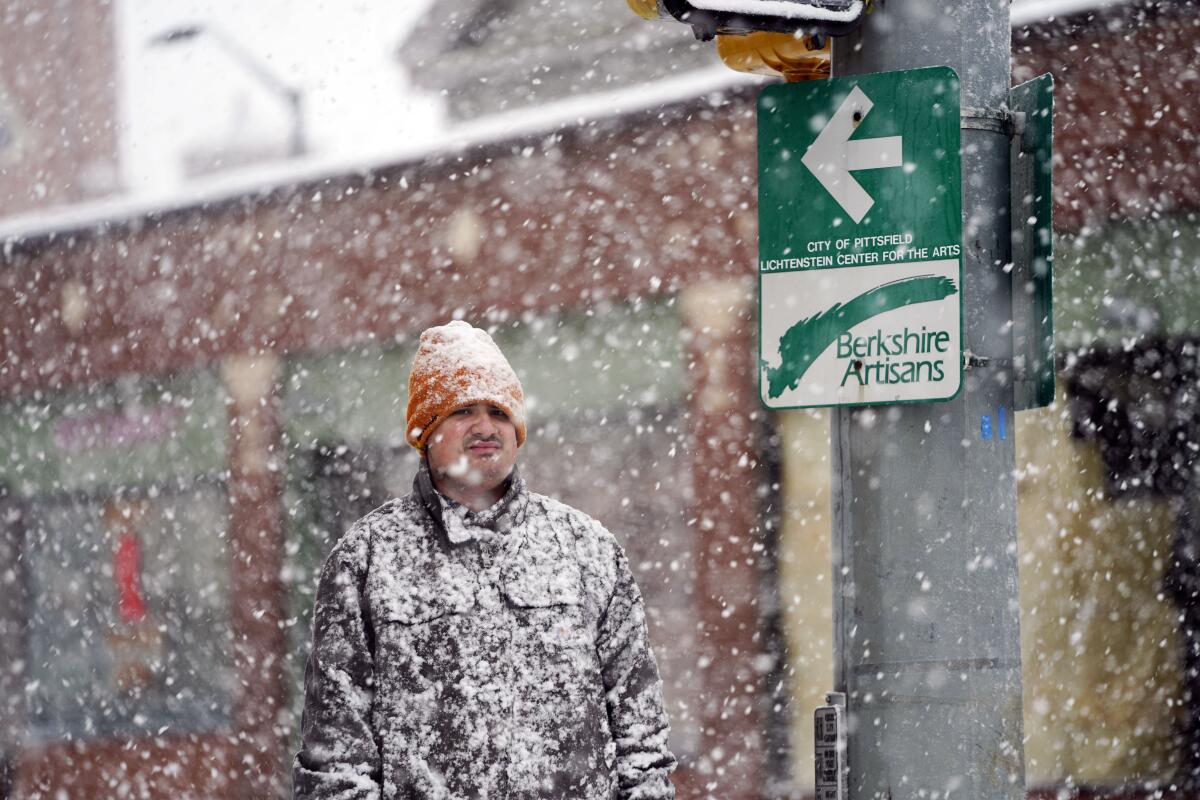 Snow falls on a man standing by a street.