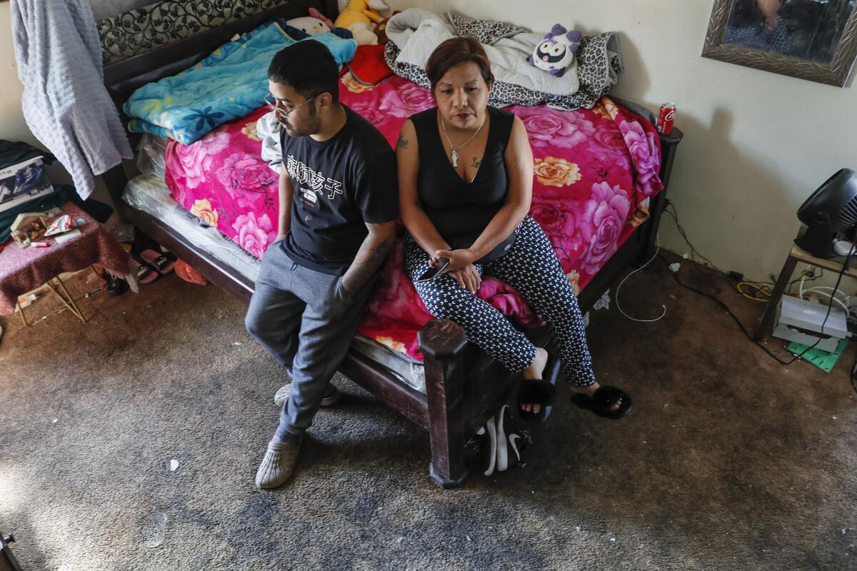 A woman and her adult son sit on a bed in a room with filthy carpeting