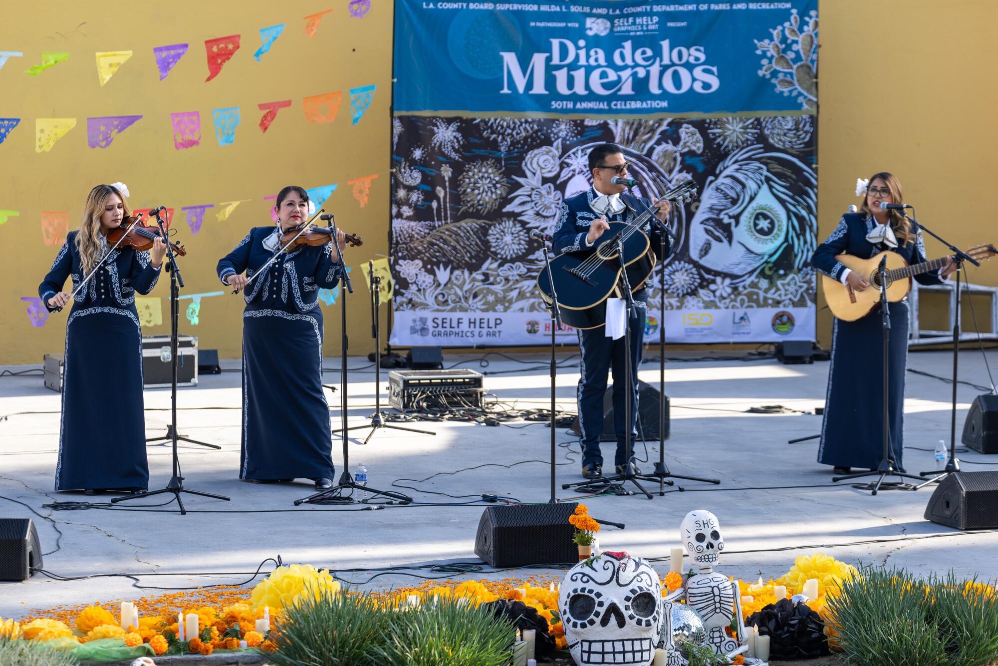 A mariachi band performs onstage. A banner behind them reads "Dia de los Muertos"