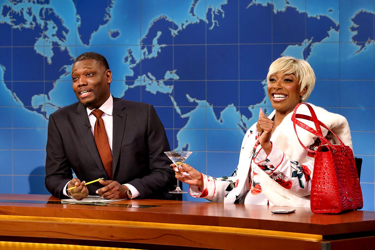 Actors Michael Che and Ego Nwodim, wearing fur and a blond wig, sit at an anchor desk set in an "SNL" skit.