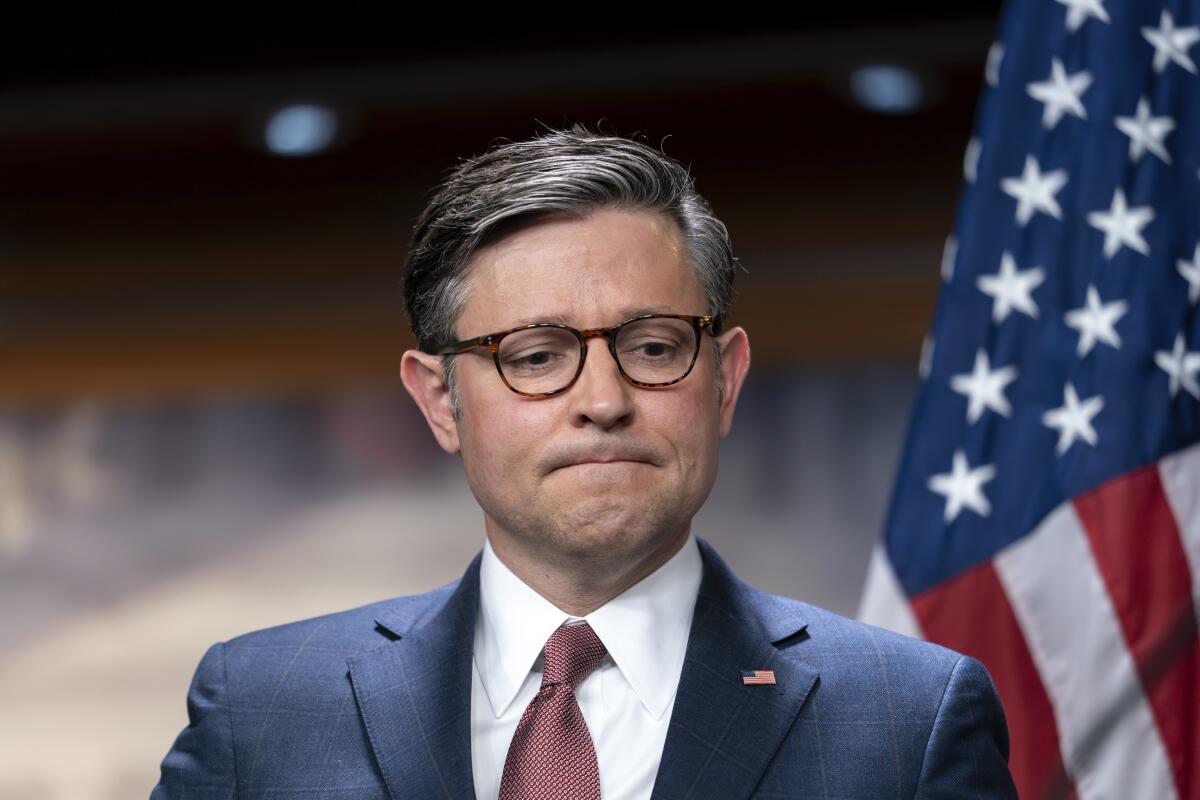 A man in a suit and glasses, looking frustrated, stands next to an American flag.