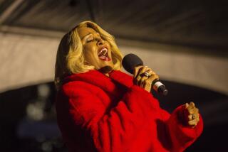 An older woman in a furry red coat sings into a microphone at a holiday event