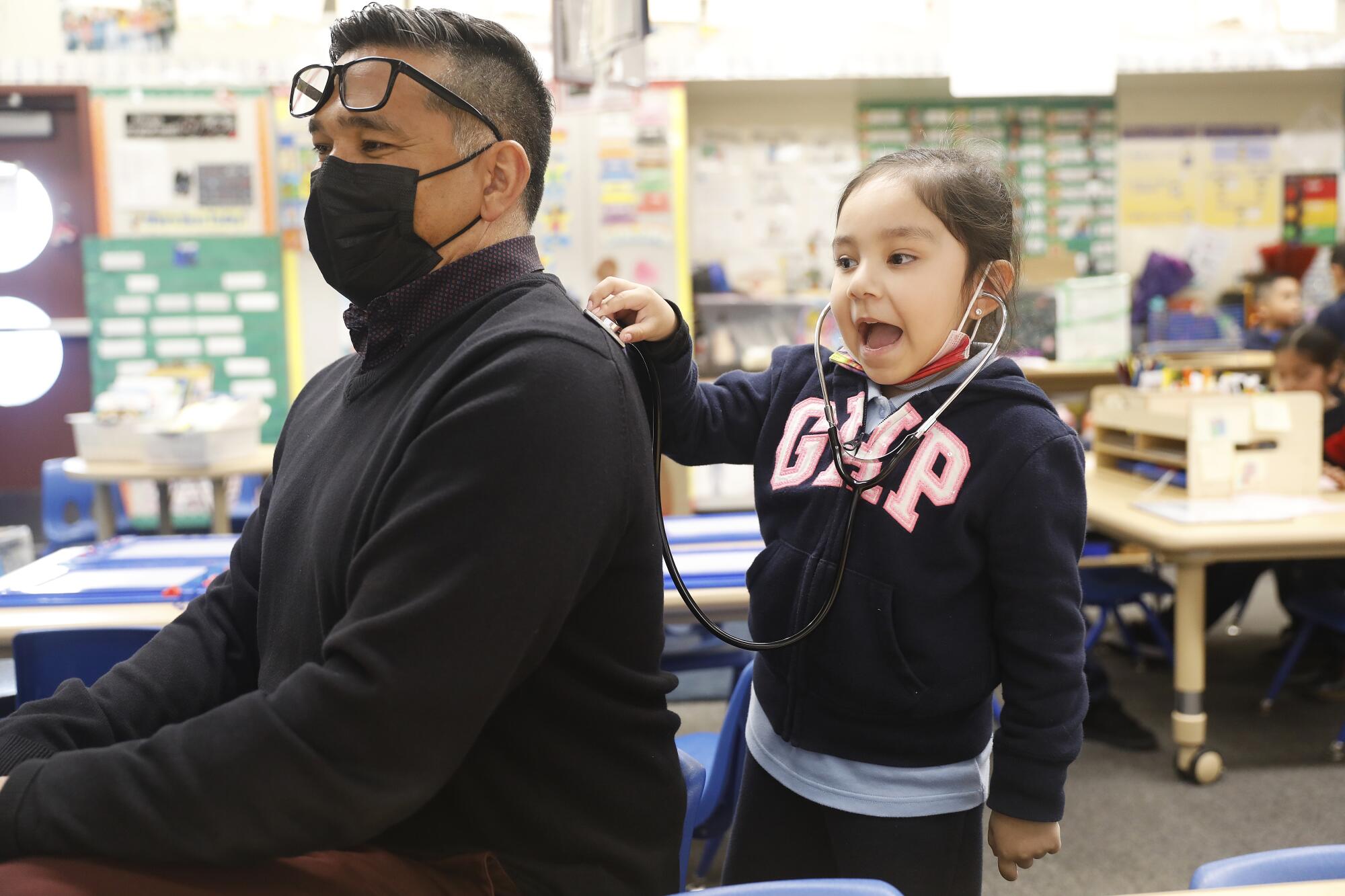 A young girl holds a stethoscope to the back of a seated adult.