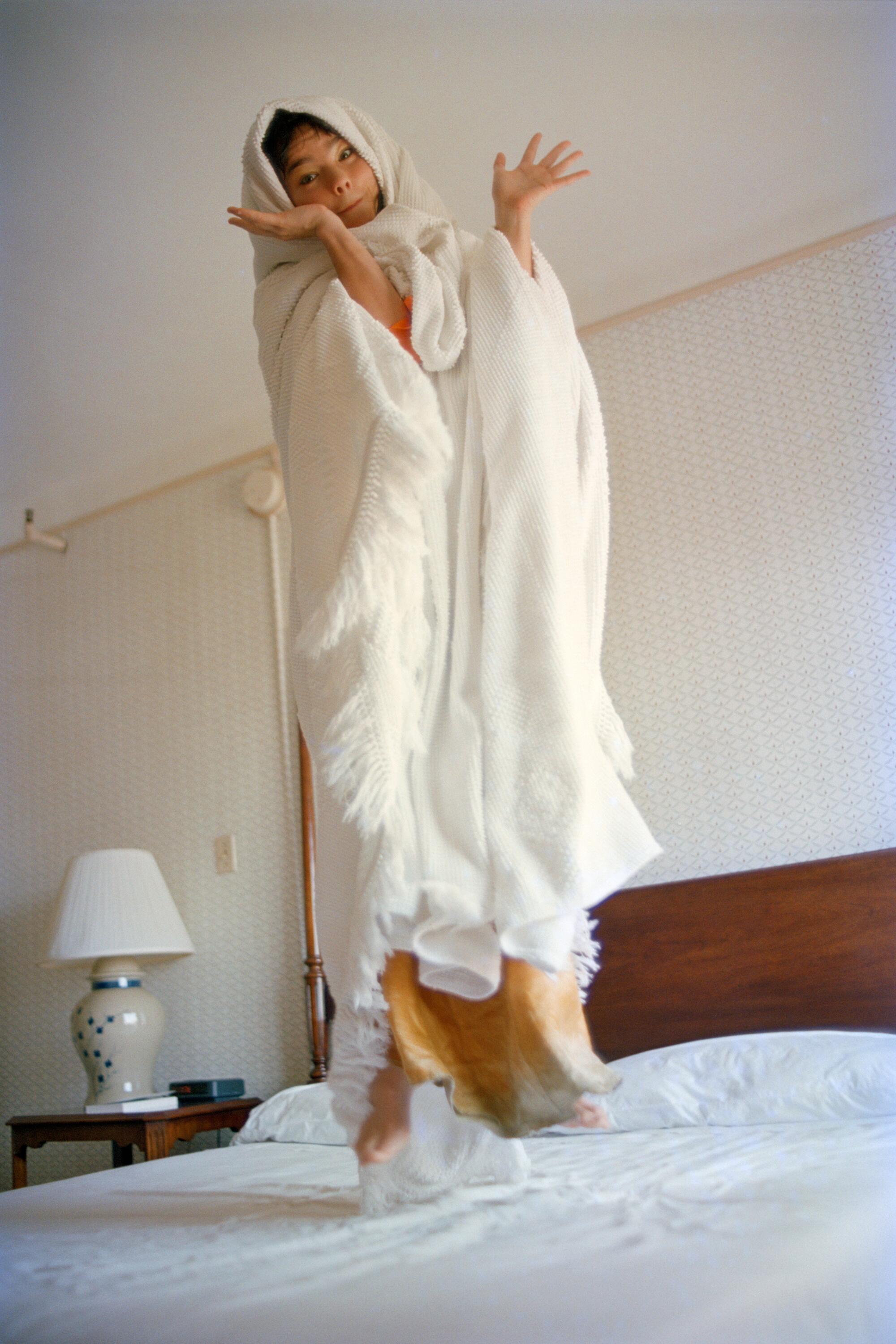 Bjrk jumps on a bed, wrapped in a white sheet, making a playful face.