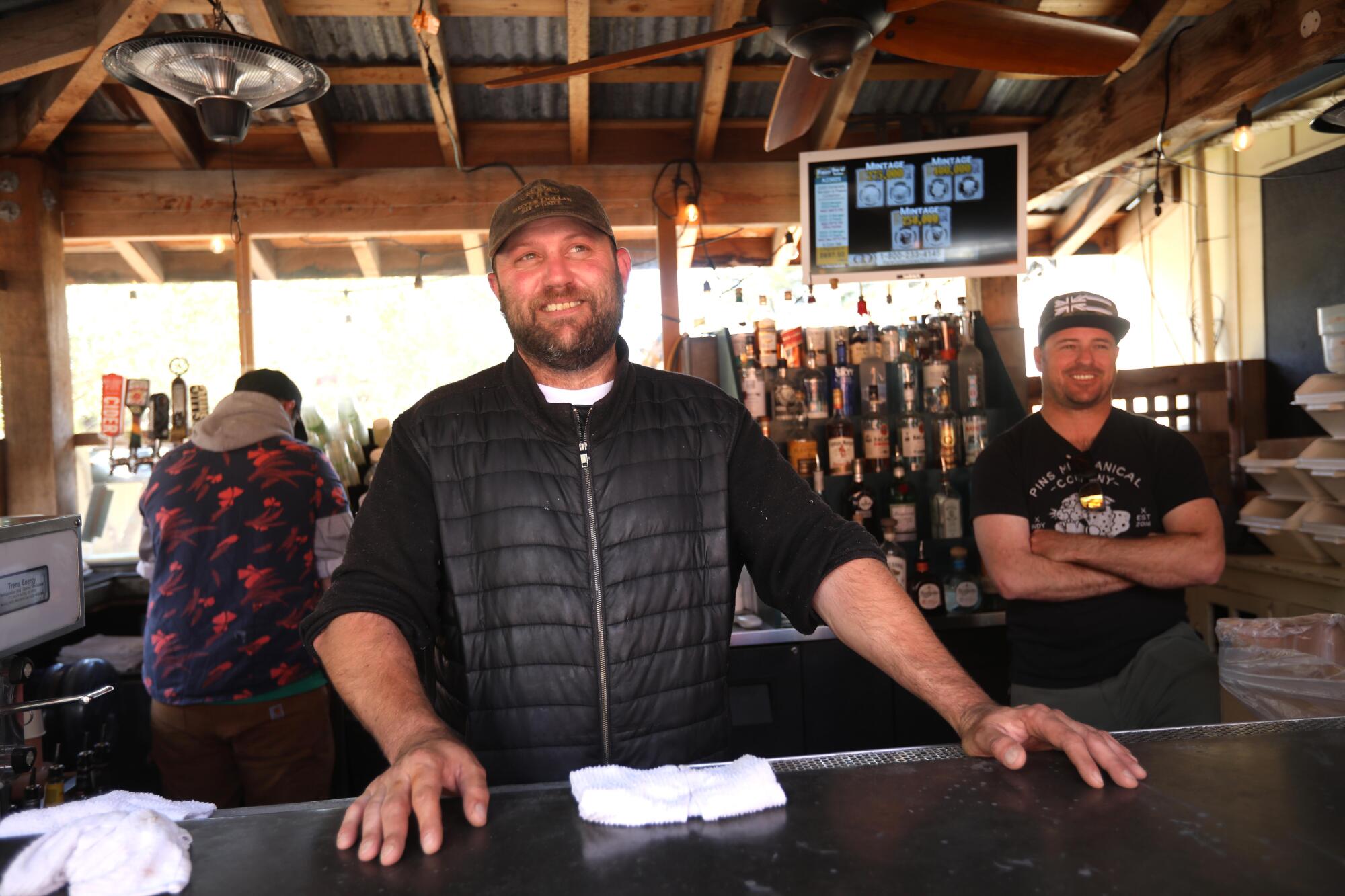 A bearded man in a down vest stands behind a bar.