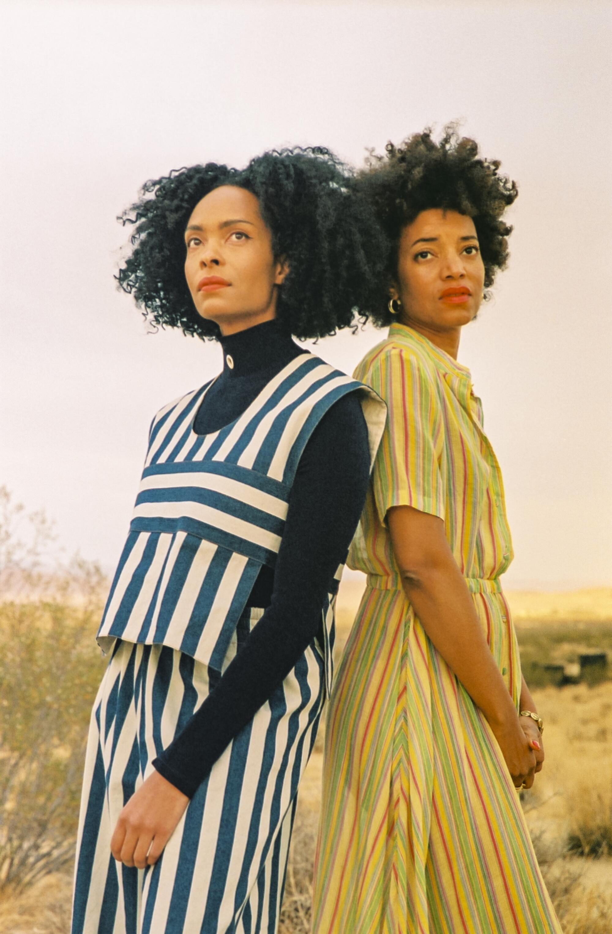 Two women stand back to back in the desert.
