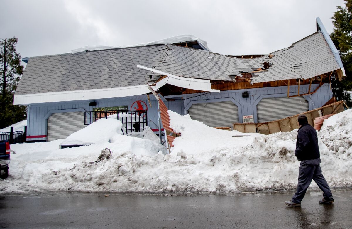A man walks past a building with a collapsed roof. Snow is piled halfway up the walls.