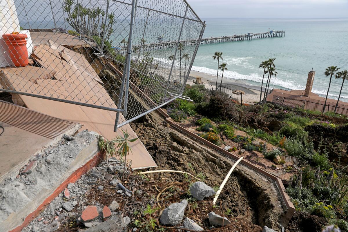 A chain-link fence teeters at the top of a crumbling bluff above palm trees and the ocean.