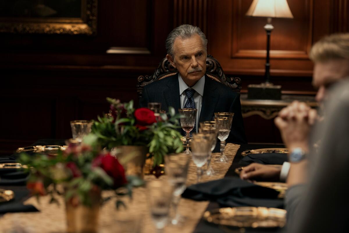An older man in a suit sits at the head of a long dinner table