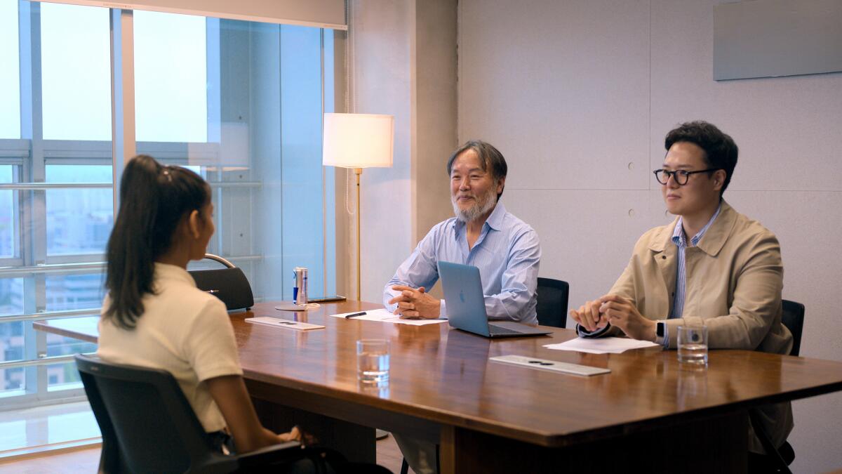 Two men sit at a desk in front of a woman.