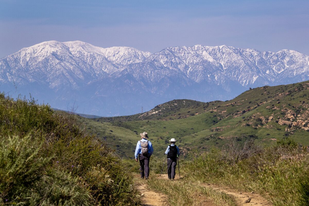 Two hikers on a lush trail in the foreground, with snow-capped mountains beyond