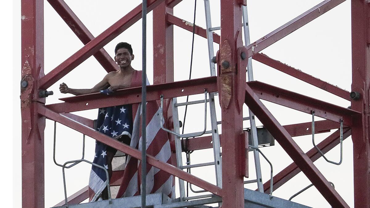 A man holds emergency personnel at bay as he descends a construction crane carrying a U.S. flag.