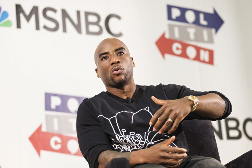 Charlamagne Tha God in a black shirt sitting down on a stage wit his hands infant of him