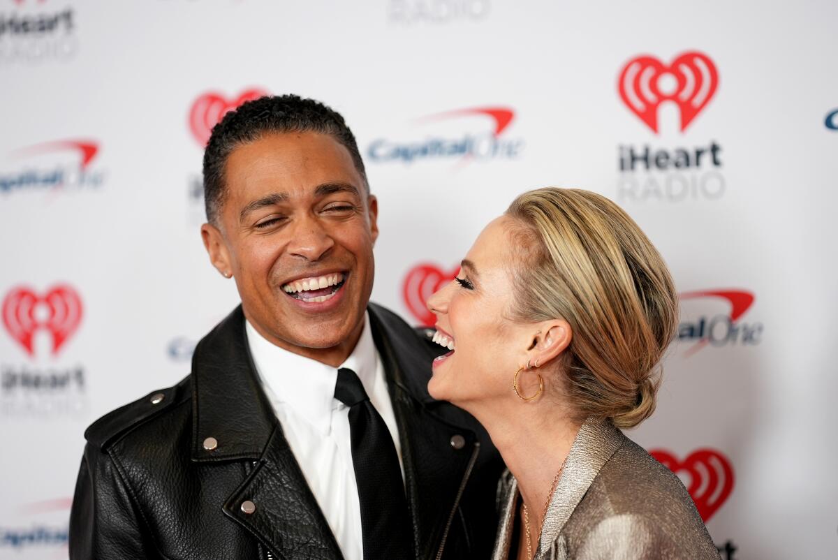 T.J. Holmes in a black jacket and white shirt laughs along with Amy Robach, clad in a gold jacket