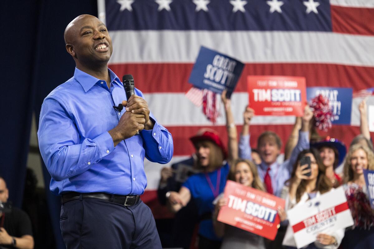 Sen. Tim Scott holds a microphone while standing before people holding signs and a U.S. flag