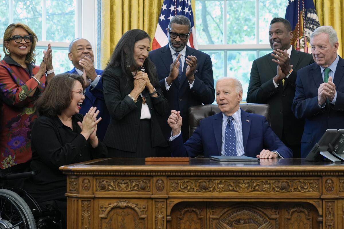 President Biden is surrounded by people  in the Oval Office.