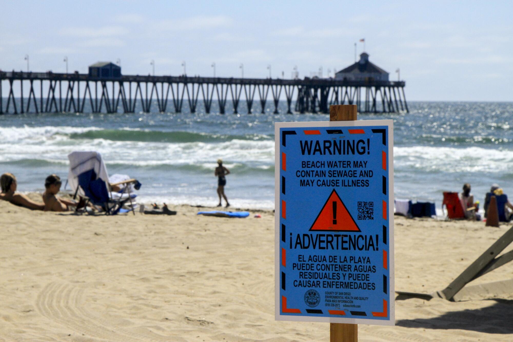 A sewage "warning" sign posted on a beach near a pier.