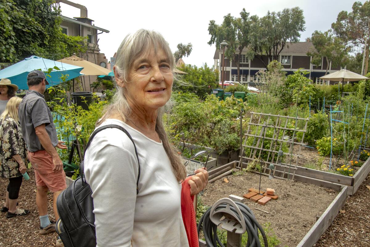 A woman in a white T-shirt with a backpack stands in a garden with raised beds.