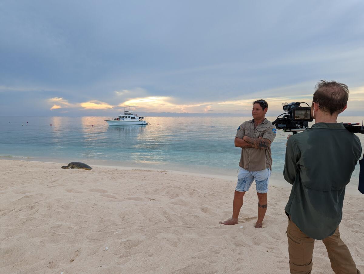 A man in a shirt and jean shorts stands on the beach as he's interviewed by a man holding a camera.