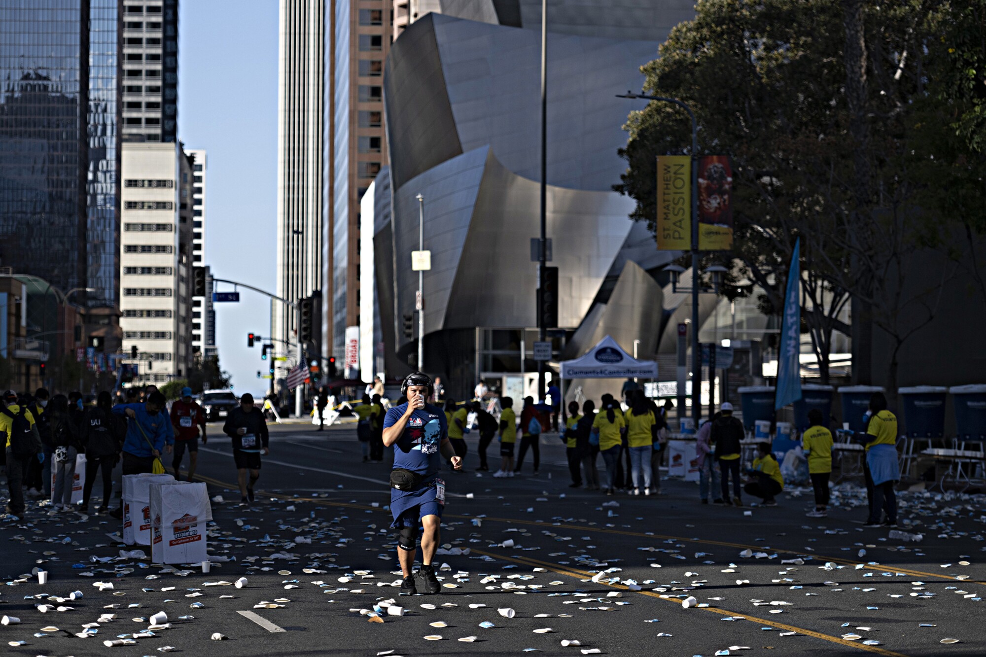 A runner drinks from a cup while running through a street littered with discarded cups with buildings in the background.