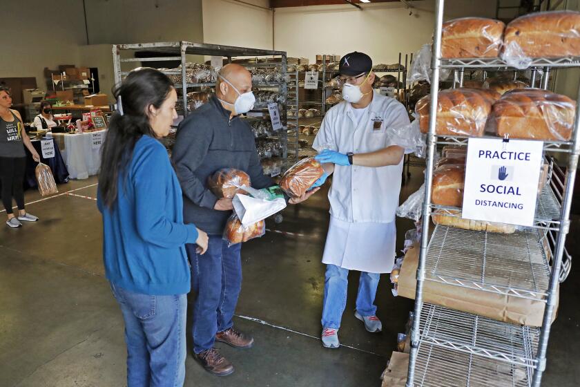 Dean Kim, right, OC Baking Company founder and artisan baker, hands Orange residents Richard Caballero and his wife Diane a loafs of bread at a grocery store pop-up that recently opened at the OC Baking Company in Orange. Kim supplies bread for a lot of hotels and restaurants in Orange County, including Toast in Newport Beach and Global Hall Eatery in Costa Mesa. With help from his community partners, they opened a pop-up grocery store that not only gives free food to health care workers but also provides a one-stop shop where locals can get food from these restaurants.