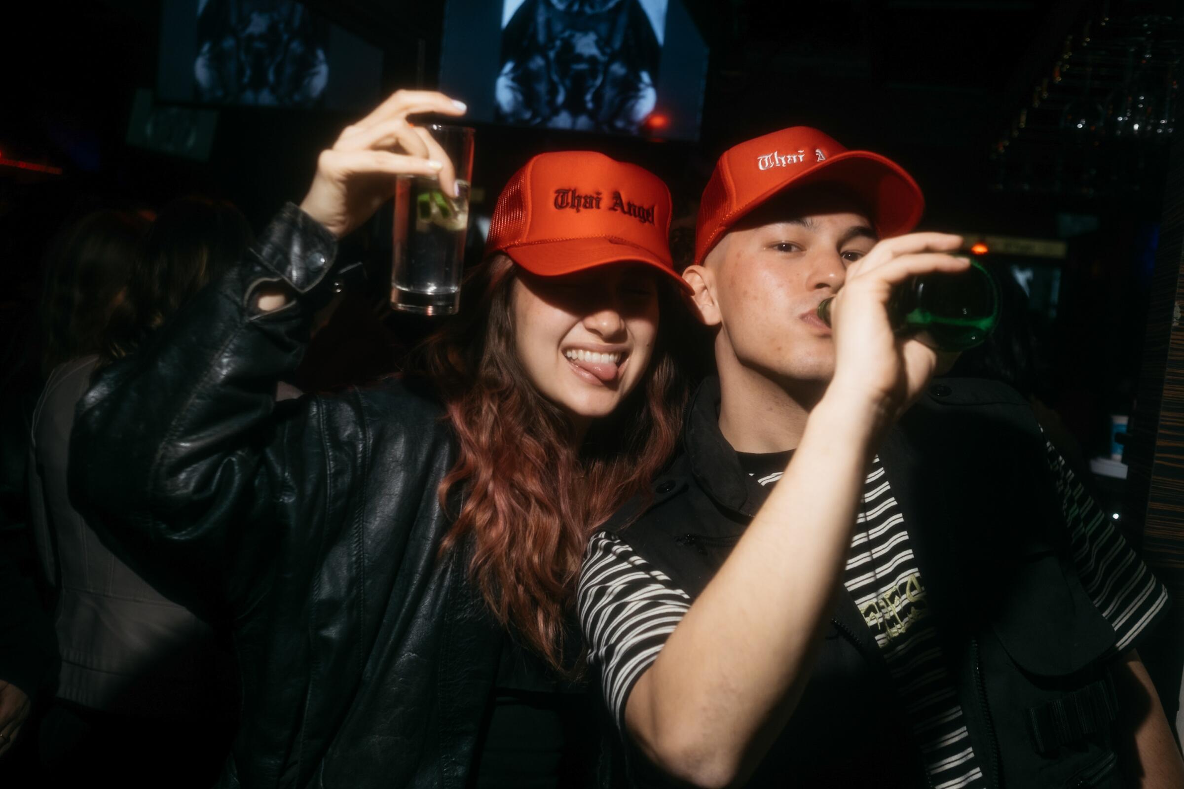 A couple wears red trucker hats that say "Thai Angel" as one drinks from a beer bottle and another holds their drink up.
